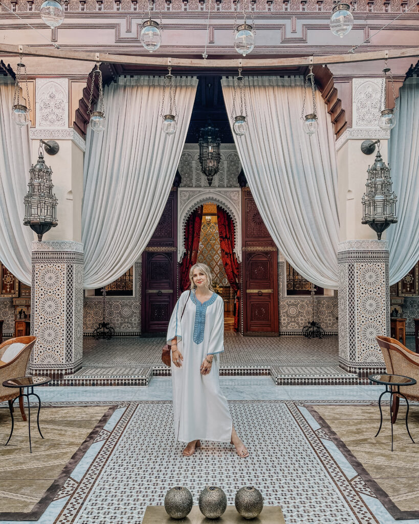 what to wear in morocco as a female traveller - blonde woman in jellaba standing in a beautiful moroccan style archway with curtains and a breeze blowing