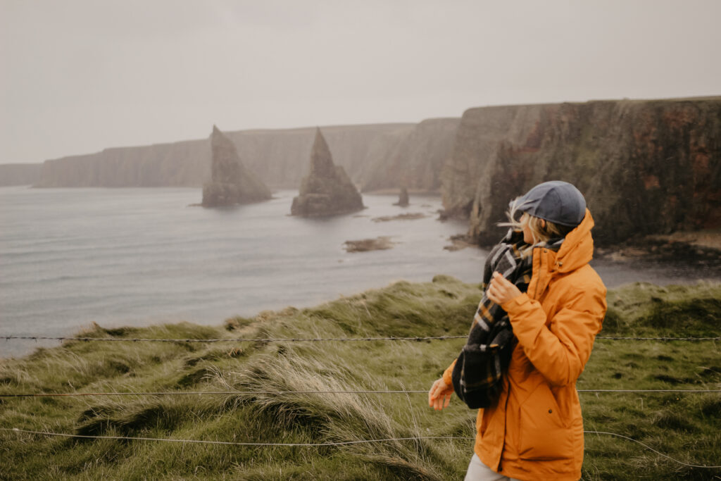 Scotland Travel Guide - Girl standing on cliffside in yellow jacket looking at the duncansby stacks.